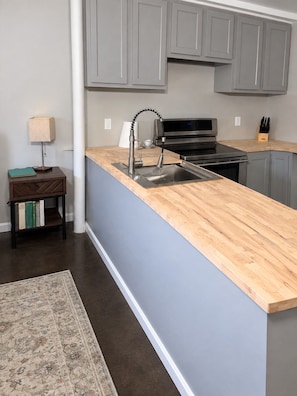 A kitchen with grey cabinets, a wooden countertop, a stainless steel sink, a stove, and a small table with a lamp and books. - Euclid Escape-Cozy Studio: Pet friendly, Free Parking, Close to Downtown (Chattanooga)