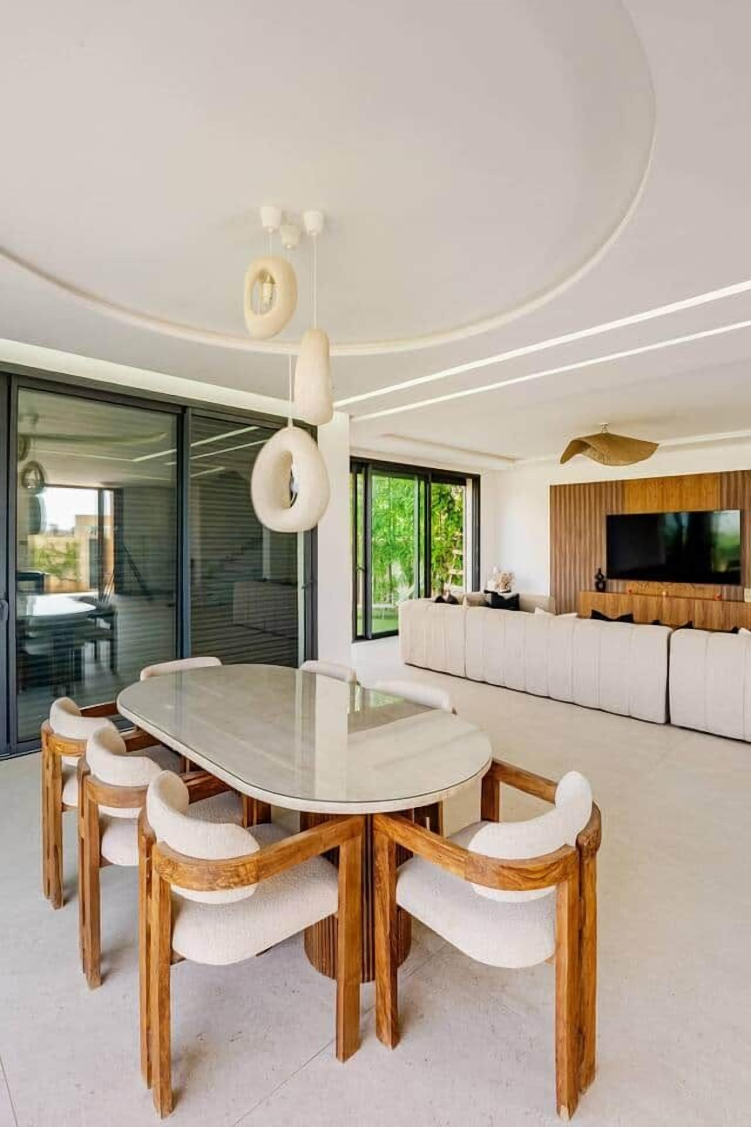 Un'area pranzo moderna con un tavolo rotondo e sei sedie, un grande televisore montato su un pannello di legno e una porta vetrata che dà su uno spazio esterno.