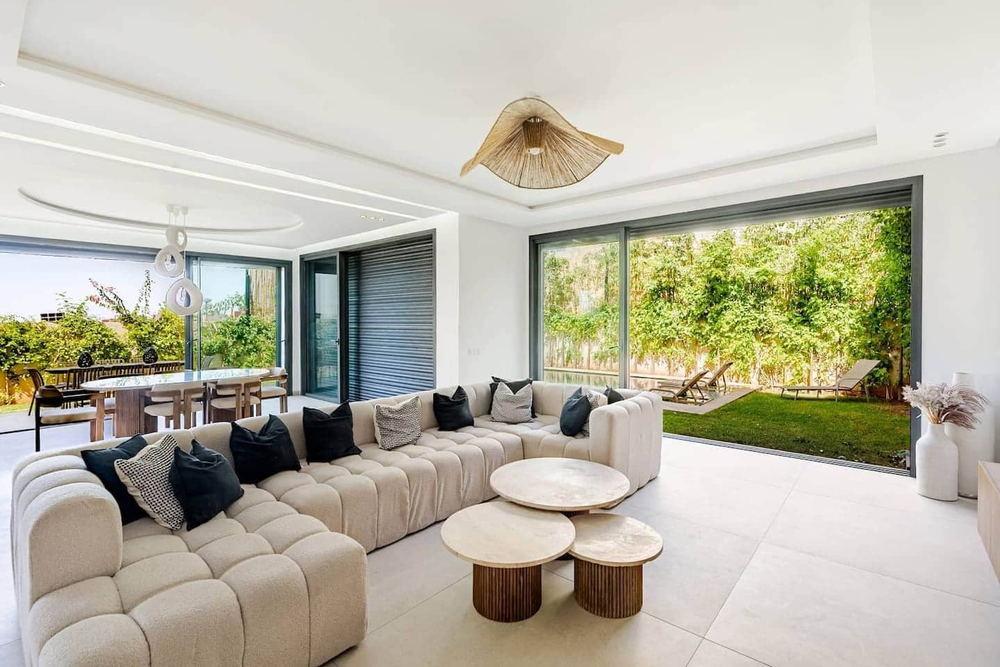 Una sala moderna con un sofá seccional, mesas de centro redondas y amplias ventanas con vistas a un jardín.
