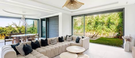 Een moderne woonkamer met een modulaire bank, ronde salontafels en grote ramen met uitzicht op een tuin.