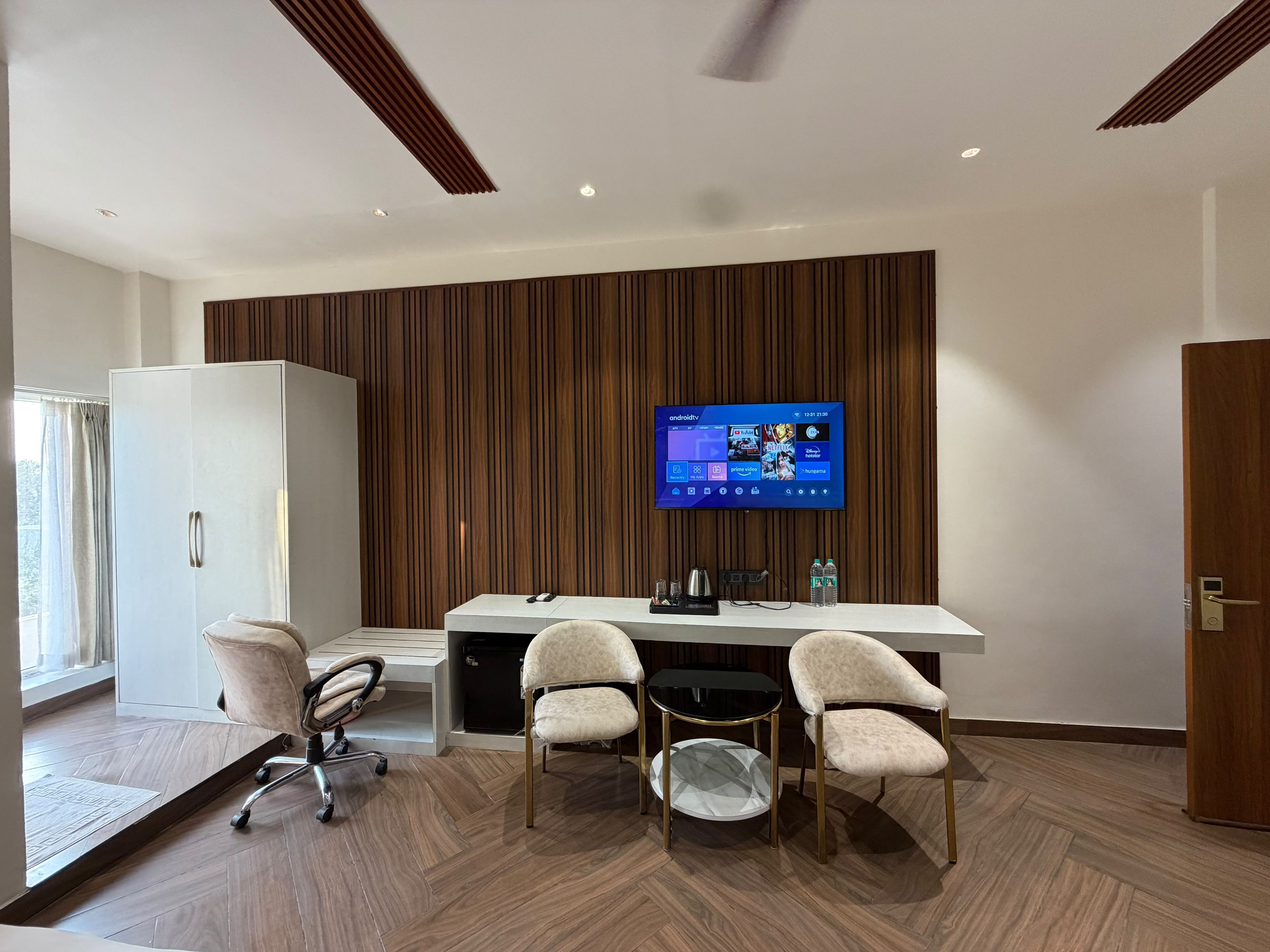 A modern conference room with a large flat-screen TV mounted on a wooden panel wall, a white desk with a coffee machine, and two plush chairs.
