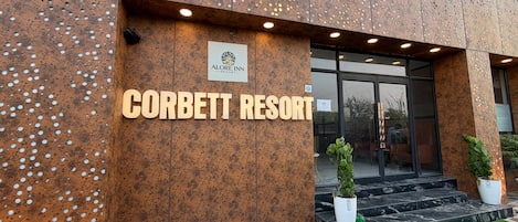 Vue extérieure du complexe Corbett Resort, avec un panneau indicateur, l’entrée et des plantes en pot.
