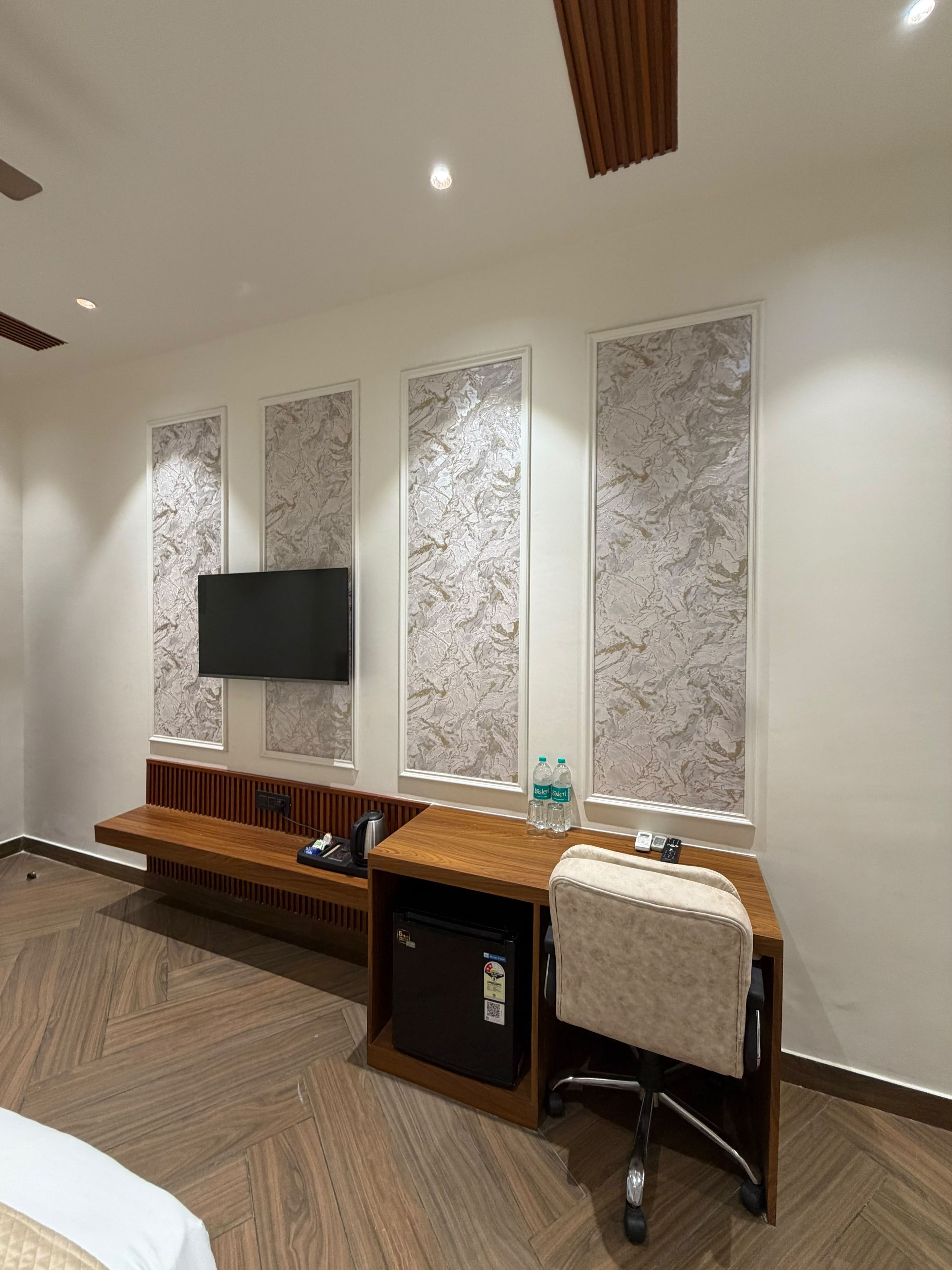 A hotel room with a wooden desk, a flat-screen TV mounted on the wall, and a seating area with a bench.