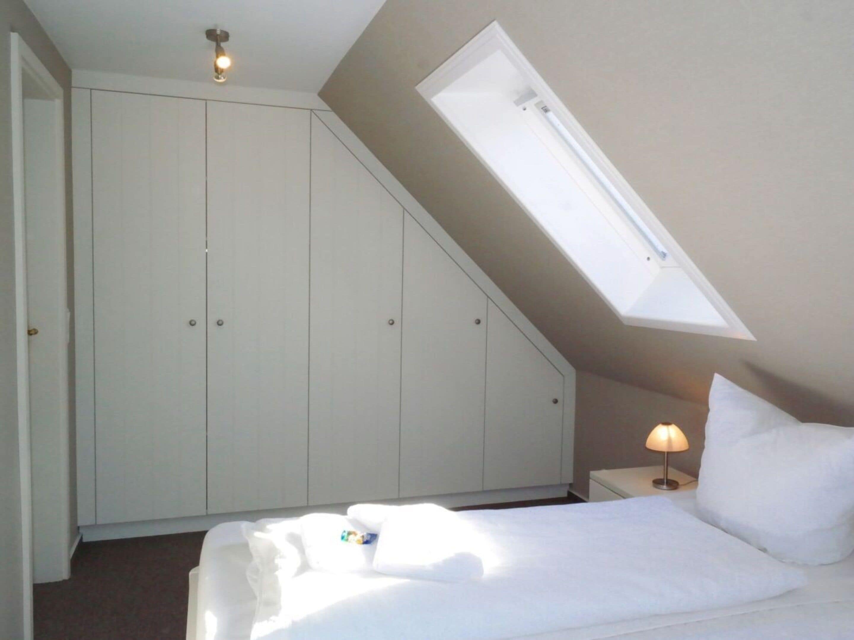 A bedroom with a skylight, a bed with white bedding, a nightstand with a lamp, and a wardrobe.