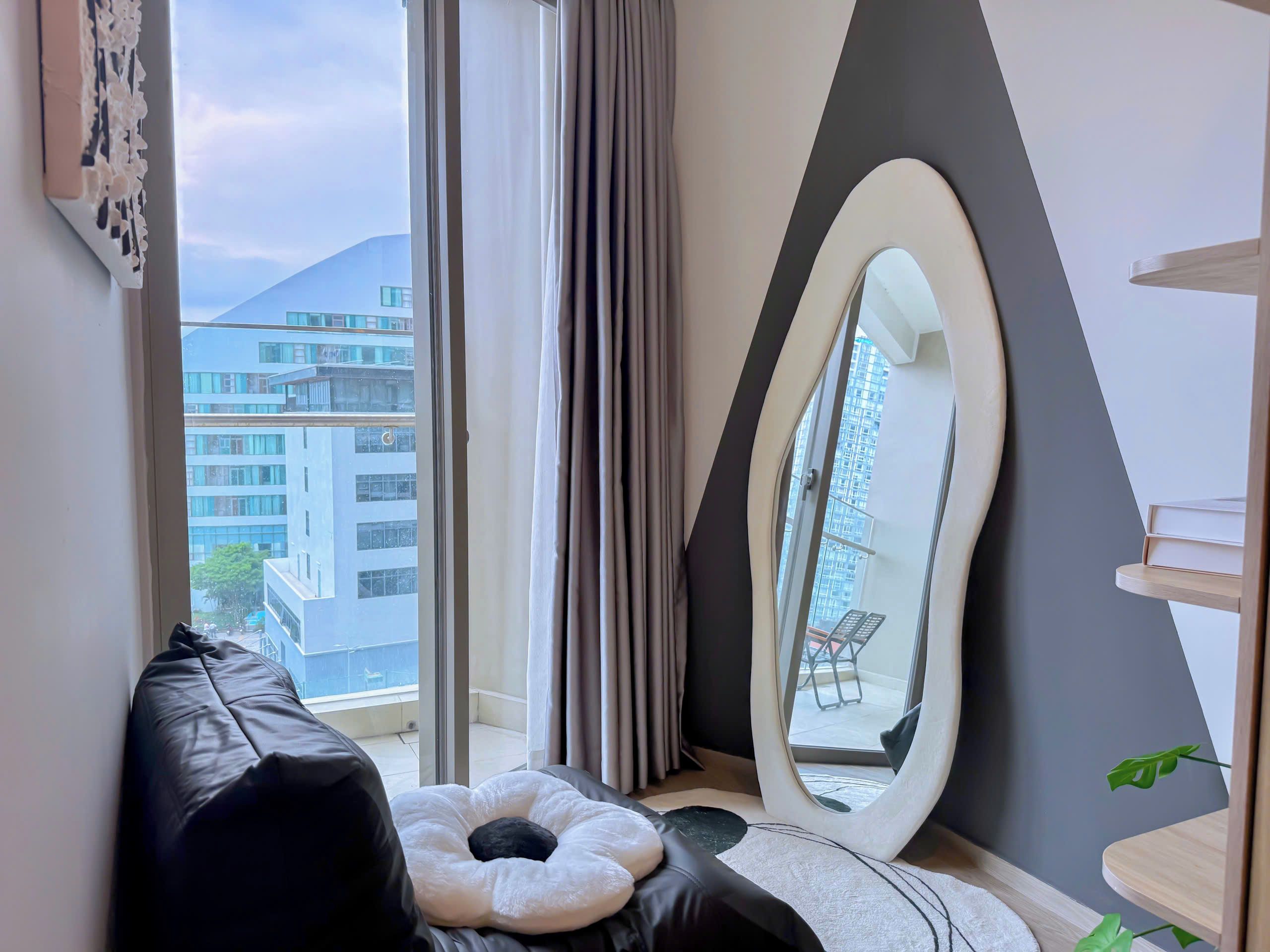 Une chambre moderne avec une grande fenêtre, un canapé en cuir noir, un coussin blanc, un miroir et une vue sur un immeuble.