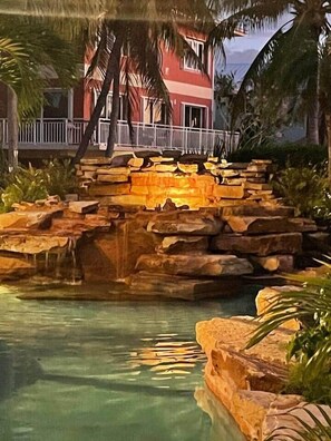 Ein Pool mit einem Felswasserfall, umgeben von tropischer Vegetation und einem Gebäude mit Balkon im Hintergrund.