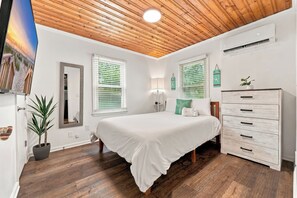 Un dormitorio con techo de madera, una cama con ropa blanca, un aparador y una planta en maceta.