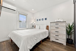 Ein Schlafzimmer mit einem Bett, einem Schminktisch, einem Nachttisch, einer Lampe, einem Fenster mit Jalousien und Wandbildern.