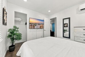 A modern bedroom with a large bed, a wall-mounted TV, a potted plant, and a beach-themed painting. - Sea Turtle at Sea Breeze Dog Friendly (North Myrtle Beach)