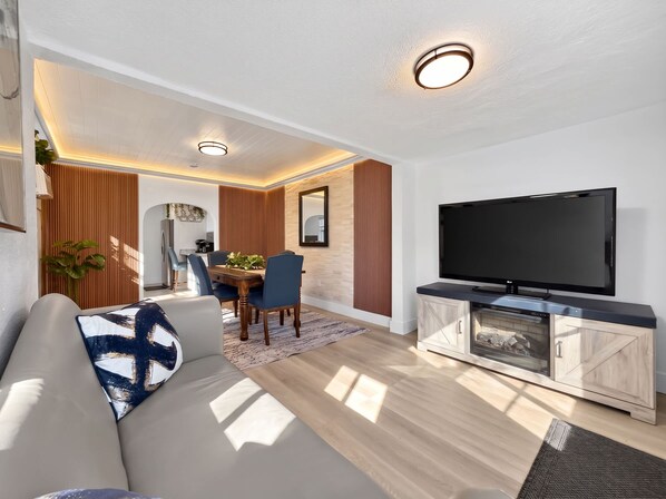 Sala de estar moderna com televisão de tela plana, sofá, área de jantar e cozinha visíveis através de um arco.