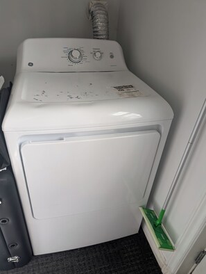 Eine Frontlader-Waschmaschine in Weiß mit Bedienelementen und einer Beschriftung, steht in einer Ecke, neben der ein Mop an der Wand lehnt.