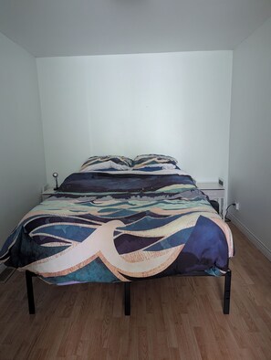 Ein Bett mit gemustertem Bettbezug und Kissen, ein Nachttisch und eine Steckdose an der Wand.