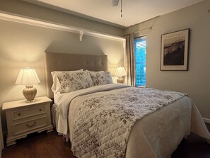 Un dormitorio con cama, dos mesitas de noche, una lámpara auxiliar, una ventana con cortinas y un cuadro colgado en la pared.