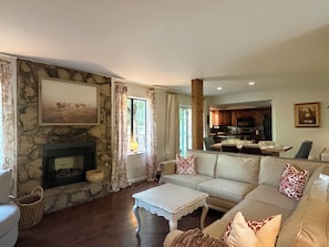 A living room with a stone fireplace, a sofa, a coffee table, and a view of the kitchen. - SEC Beauty Offers Outdoor Oasis & Entertaining! Gorgeous Yard & Privacy (Norman)