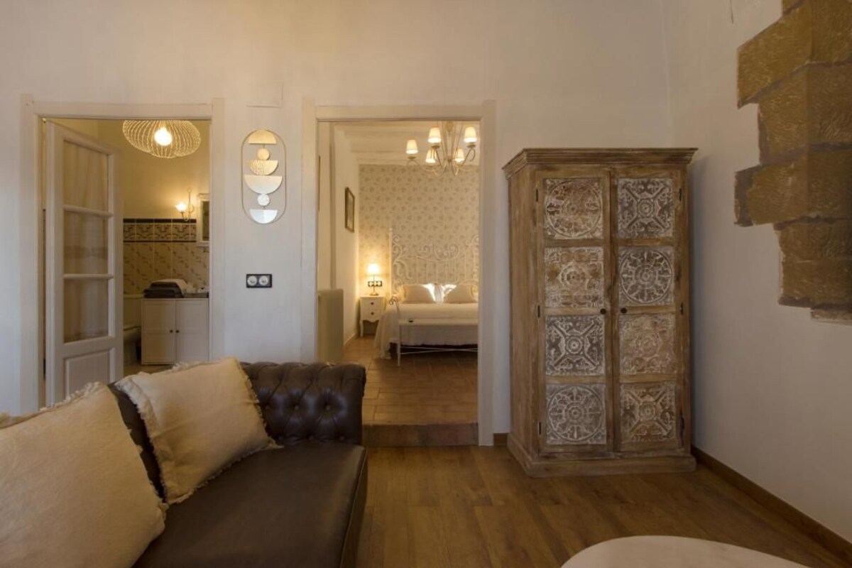 Una stanza con un divano in pelle marrone, un mobiletto in legno con decorazioni elaborate, e un'apertura che dà su un'altra stanza con un letto e un comodino.