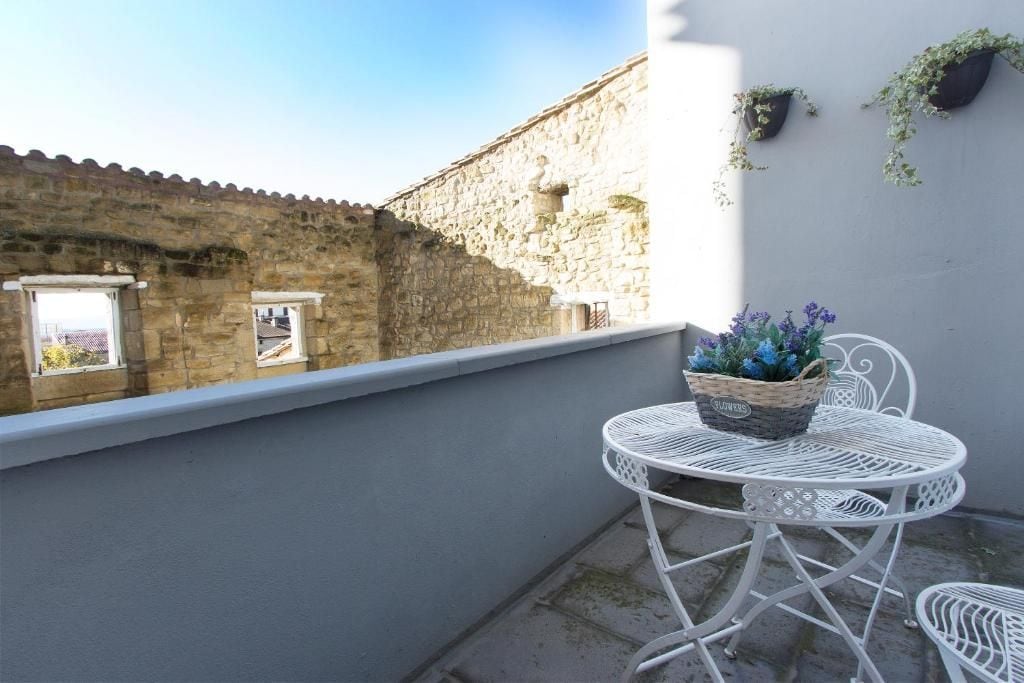 A balcony with a white metal table and chairs, a wicker basket with flowers, and a stone wall in the background.