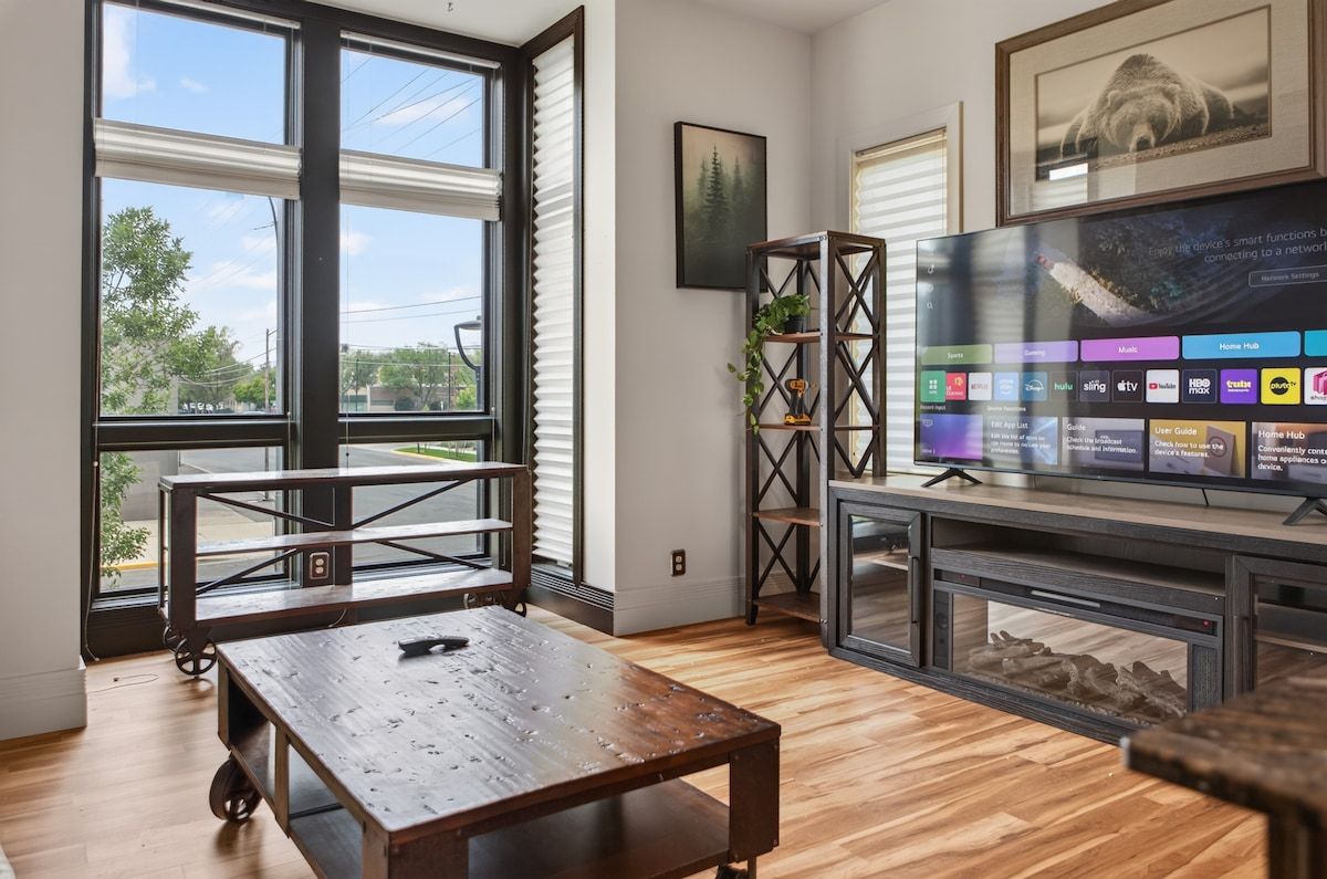 A living room with a large window, a wooden table, a TV on a stand, and a plant.