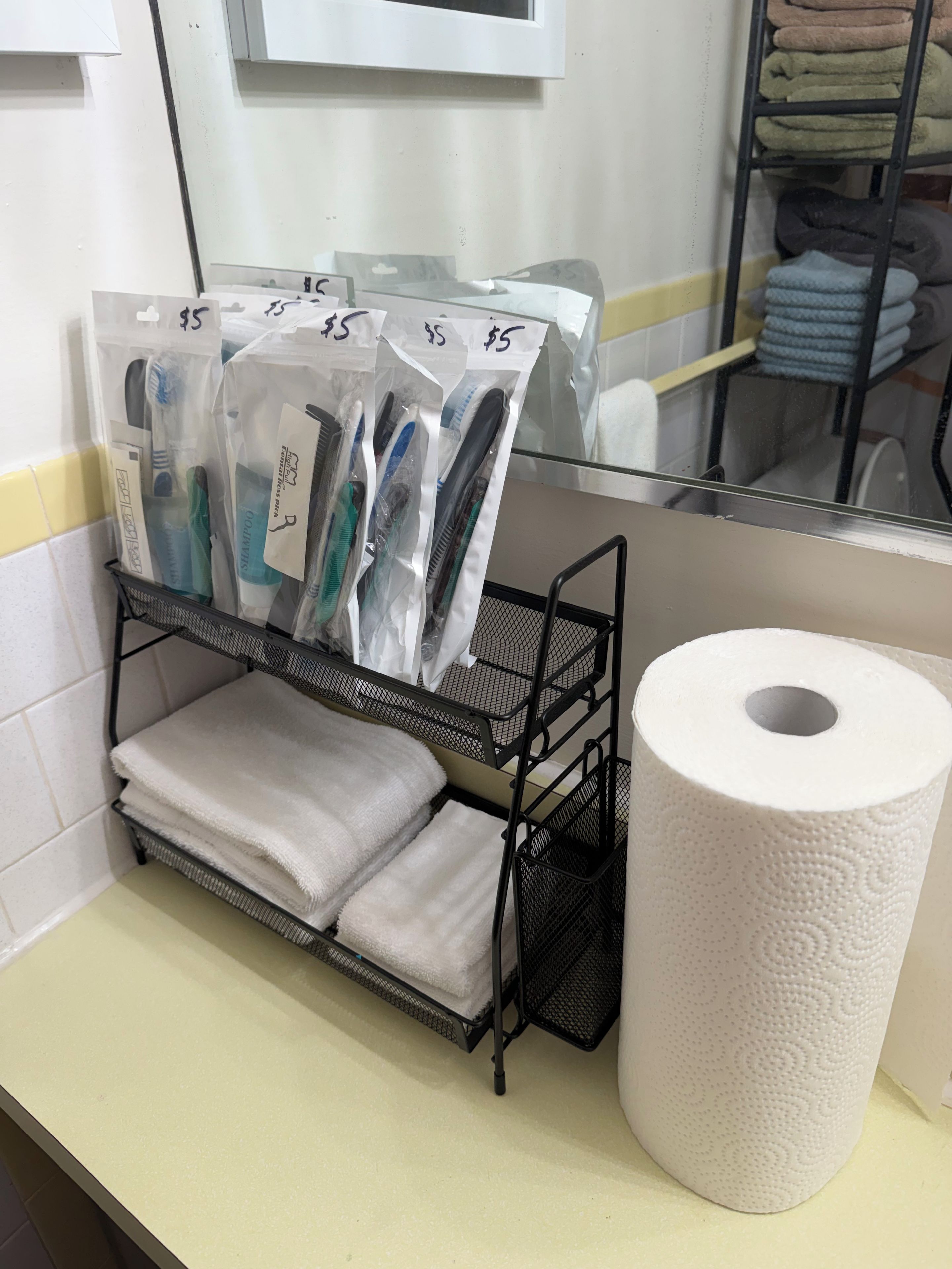 Une salle de bain avec un miroir, un porte-serviettes avec des serviettes pliées, un présentoir avec des articles de toilette dans des sacs en plastique et un rouleau de papier toilette.