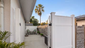 A narrow concrete pathway leading to a patio area with two chairs and a table, surrounded by a white fence and a wooden wall.