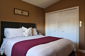 A bedroom with a bed, pillows, a bedside table, a nightstand, a framed picture on the wall, and a white door. - Melrose Heights | 4BR Executive Home (Timmins)