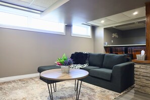 A living room with a gray sofa, a round wooden coffee table, and a potted plant with colorful flowers. - Melrose Heights | 4BR Executive Home (Timmins)