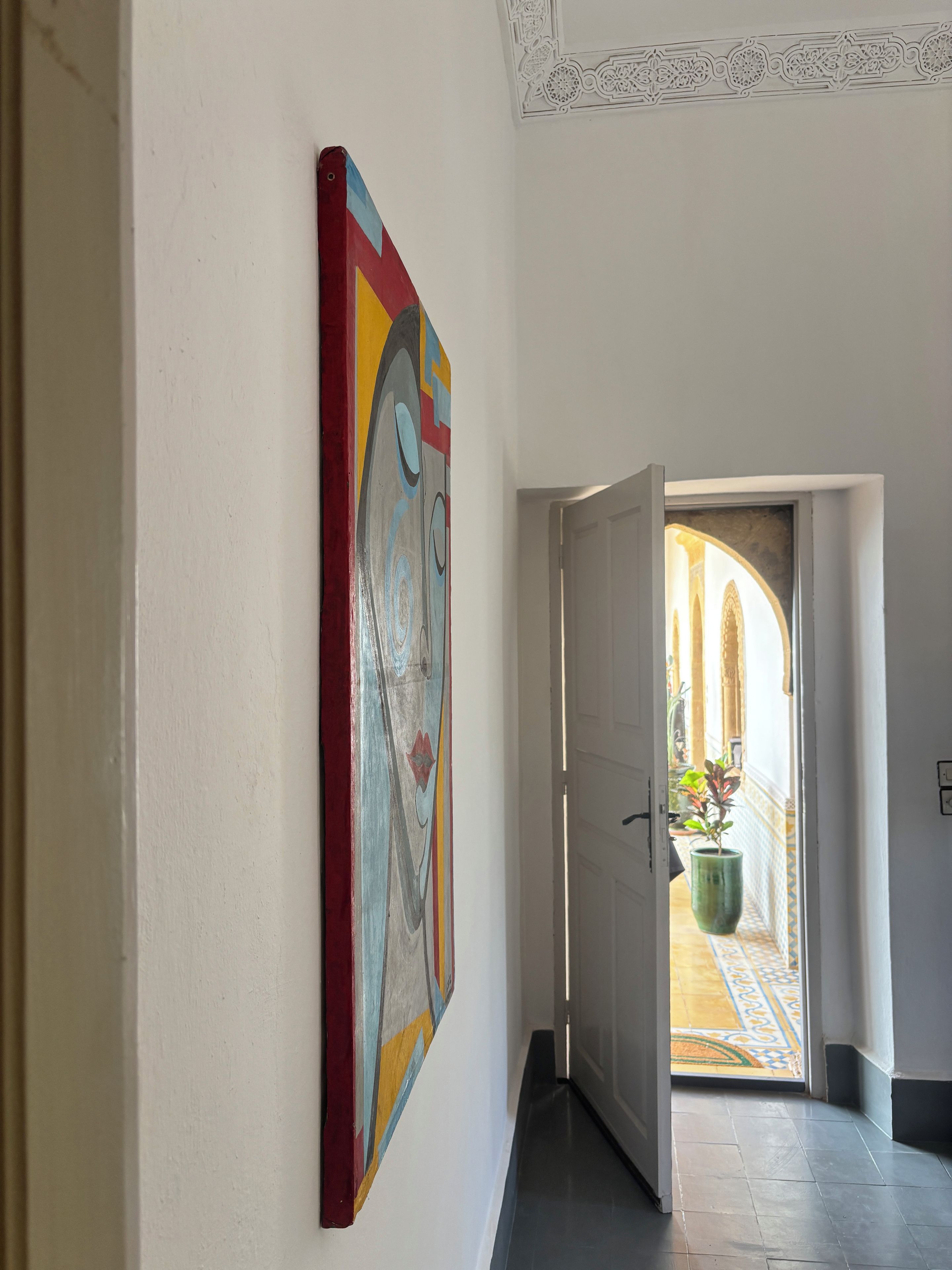 Un couloir orné d’un tableau abstrait aux couleurs vives, une porte donnant sur un espace carrelé où se trouve un pot de fleurs, et un sol à motifs.