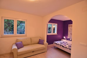 Una stanza con un divano color beige, un letto con lenzuola viola e due finestre che danno su alberi.