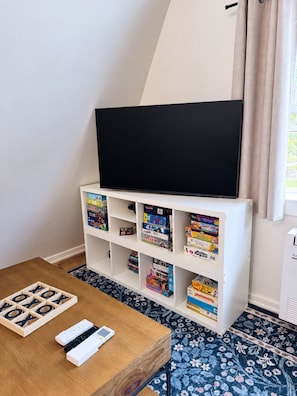 A room with a flat-screen TV on a white stand, a bookshelf with various board games, a wooden table with game pieces, and a patterned floor. - Penney Pines: A Cozy Catskills A-Frame (Catskill)