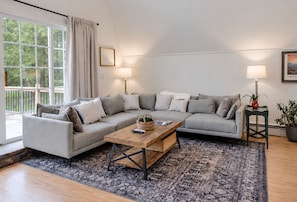 A modern living room with a grey sectional sofa, a wooden coffee table, and a patterned rug. There are two floor lamps and a potted plant on a side table. - Penney Pines: A Cozy Catskills A-Frame (Catskill)