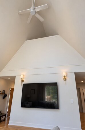 A room with a large flat-screen TV mounted on the wall, a ceiling fan, and two wall-mounted light fixtures. - Penney Pines: A Cozy Catskills A-Frame (Catskill)