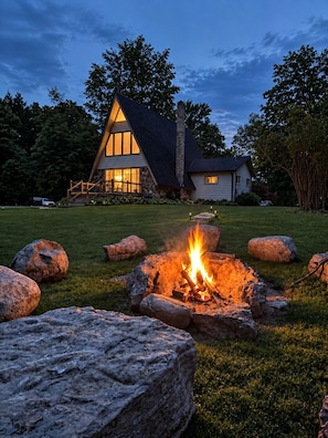 A house with a stone chimney, a fire pit with logs, and a well-maintained lawn. - Penney Pines: A Cozy Catskills A-Frame (Catskill)