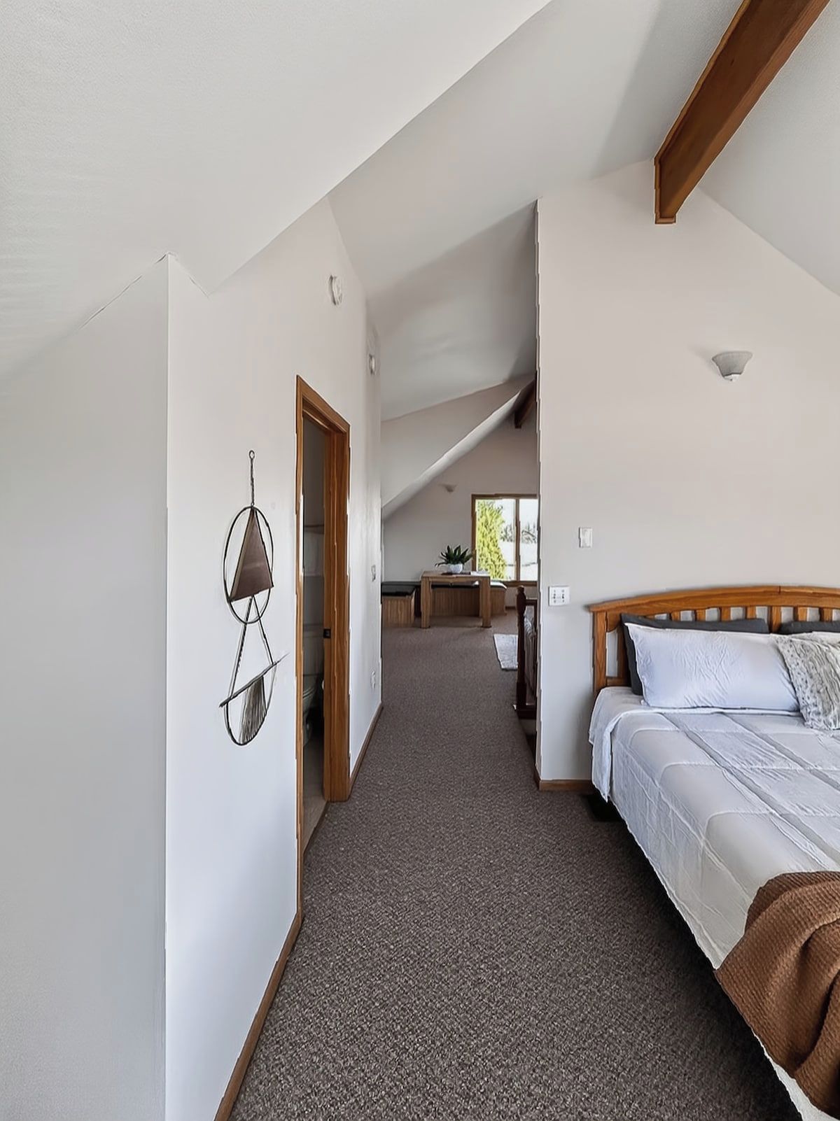 A bedroom with a wooden headboard, a bed with white bedding, a carpeted floor, and a hallway leading to a dining area.