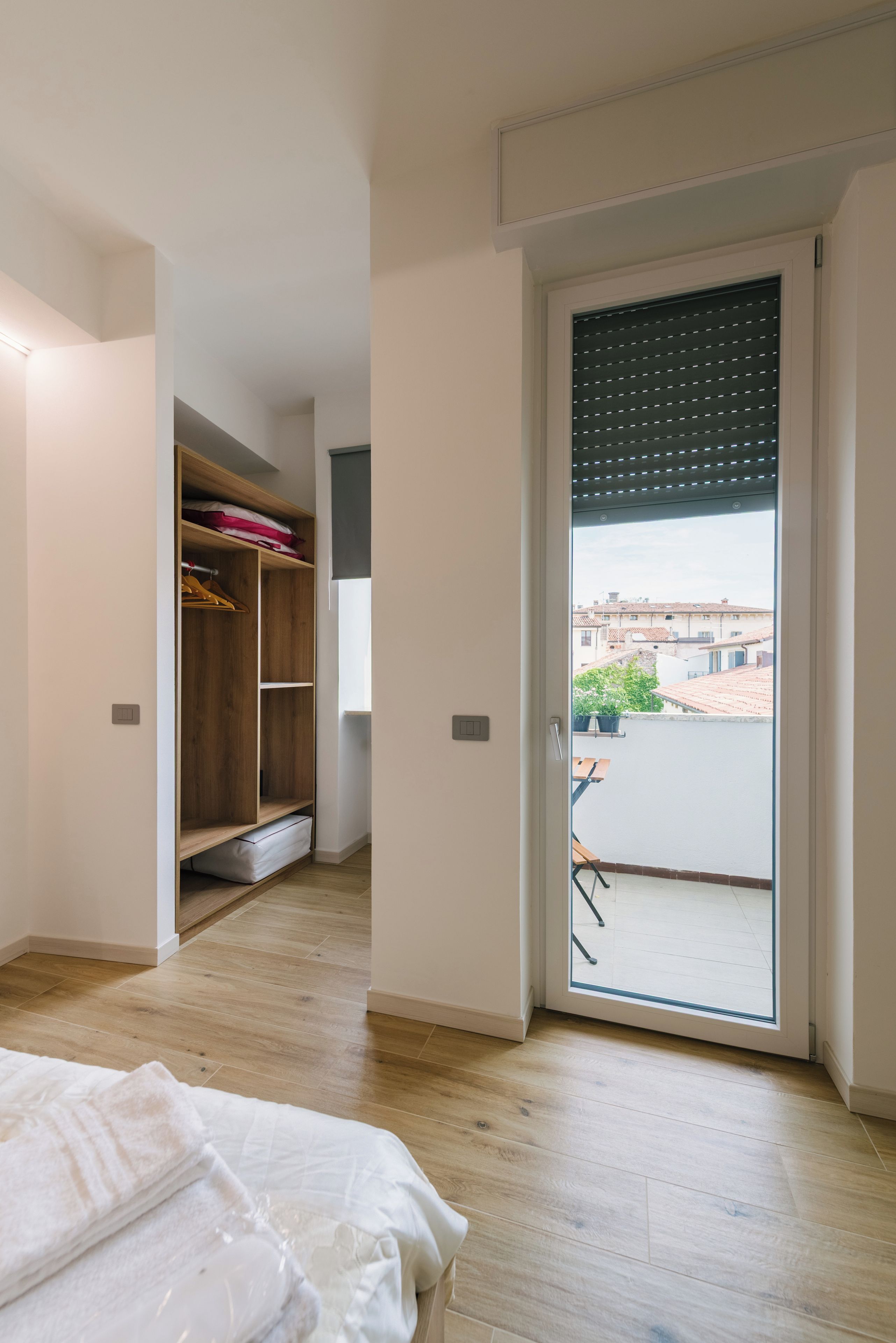 Una stanza con pavimento in legno, un letto con biancheria bianca, un armadio con ripiani in legno e una porta scorrevole in vetro che dà su una zona esterna con vista sugli edifici.