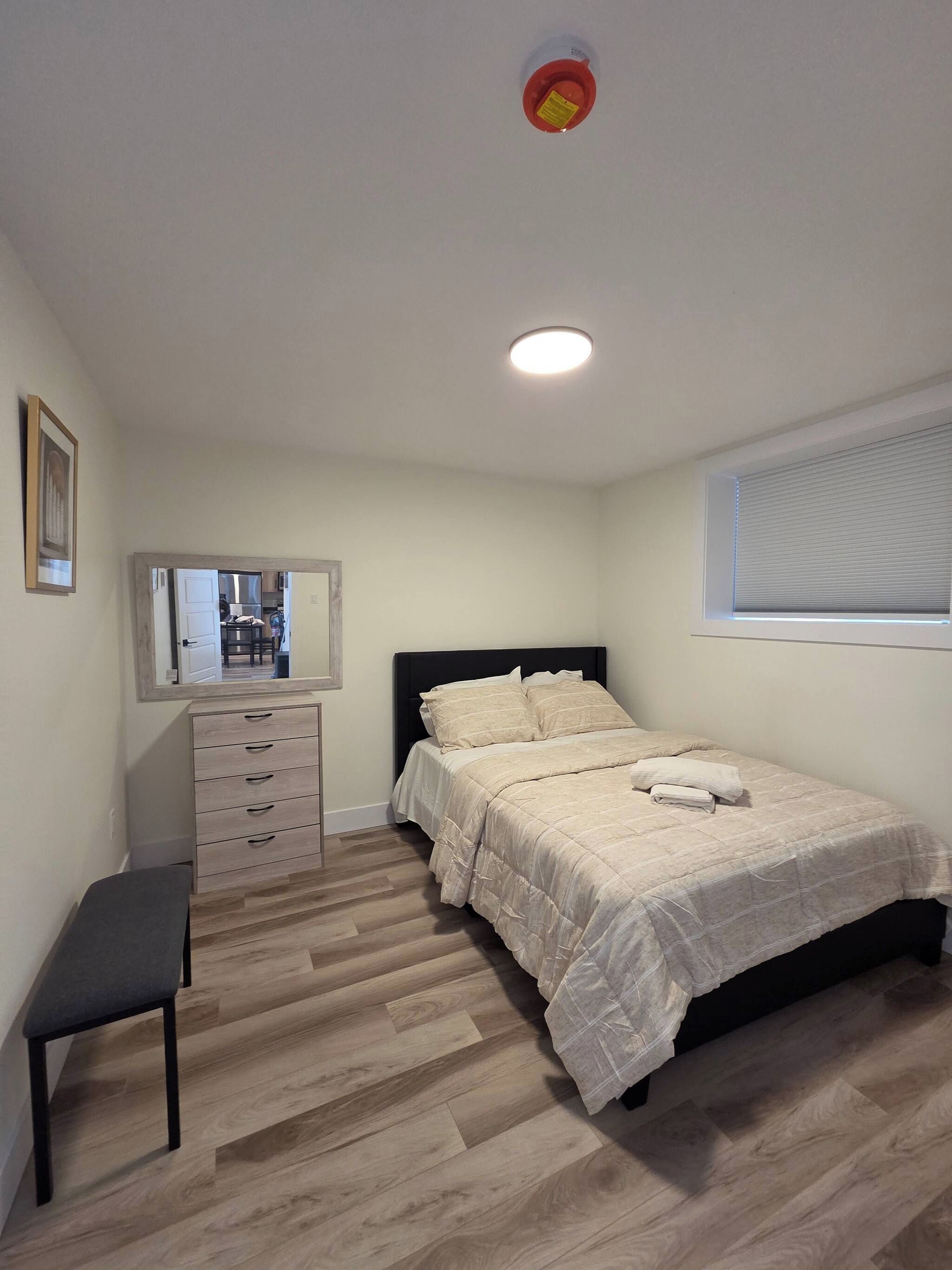 Ein Schlafzimmer mit einem Bett, einem Schminktisch, einer Bank, einem Spiegel und einem Fenster mit Jalousien.