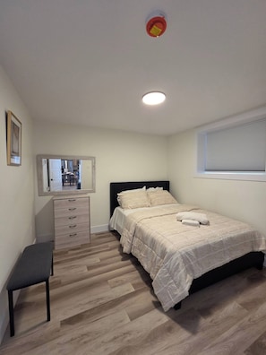 Ein Schlafzimmer mit einem Bett, einem Schminktisch, einer Bank, einem Spiegel und einem Fenster mit Jalousien.