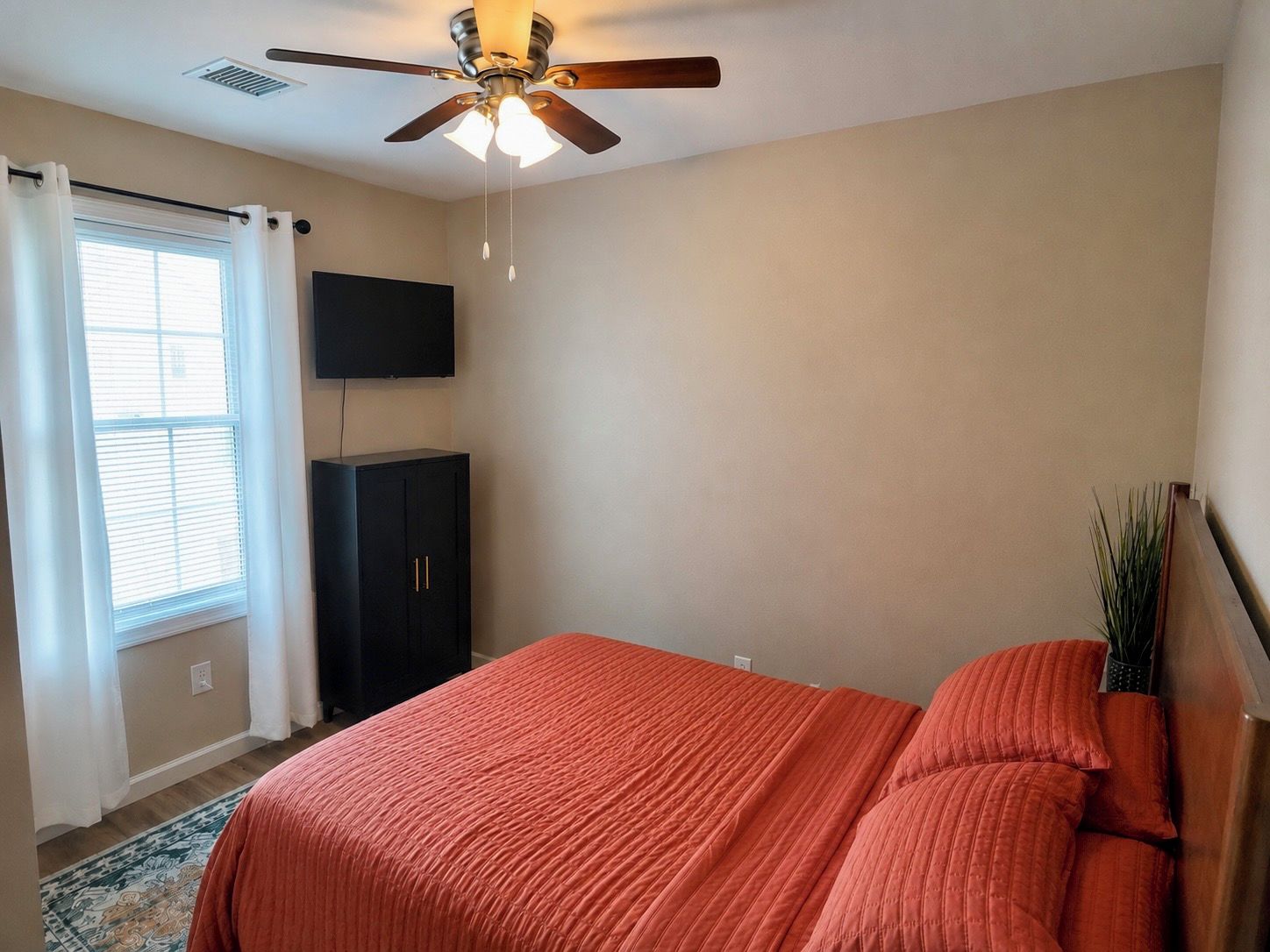A bedroom with a bed, a ceiling fan, a TV mounted on the wall, and a window with curtains.