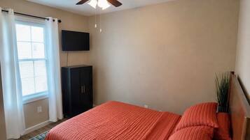 A bedroom with a bed, a ceiling fan, a TV mounted on the wall, and a window with curtains.