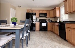 A modern kitchen with wooden cabinets, a black refrigerator, a microwave, and a stove. There is a dining table with chairs and a potted plant.