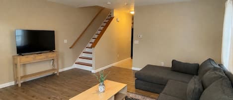 A modern living room with a flat-screen TV, a wooden coffee table, a gray sofa, a patterned rug, and a staircase with wooden steps.