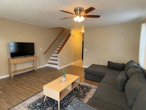 A modern living room with a flat-screen TV, a wooden coffee table, a gray sofa, a patterned rug, and a staircase with wooden steps.
