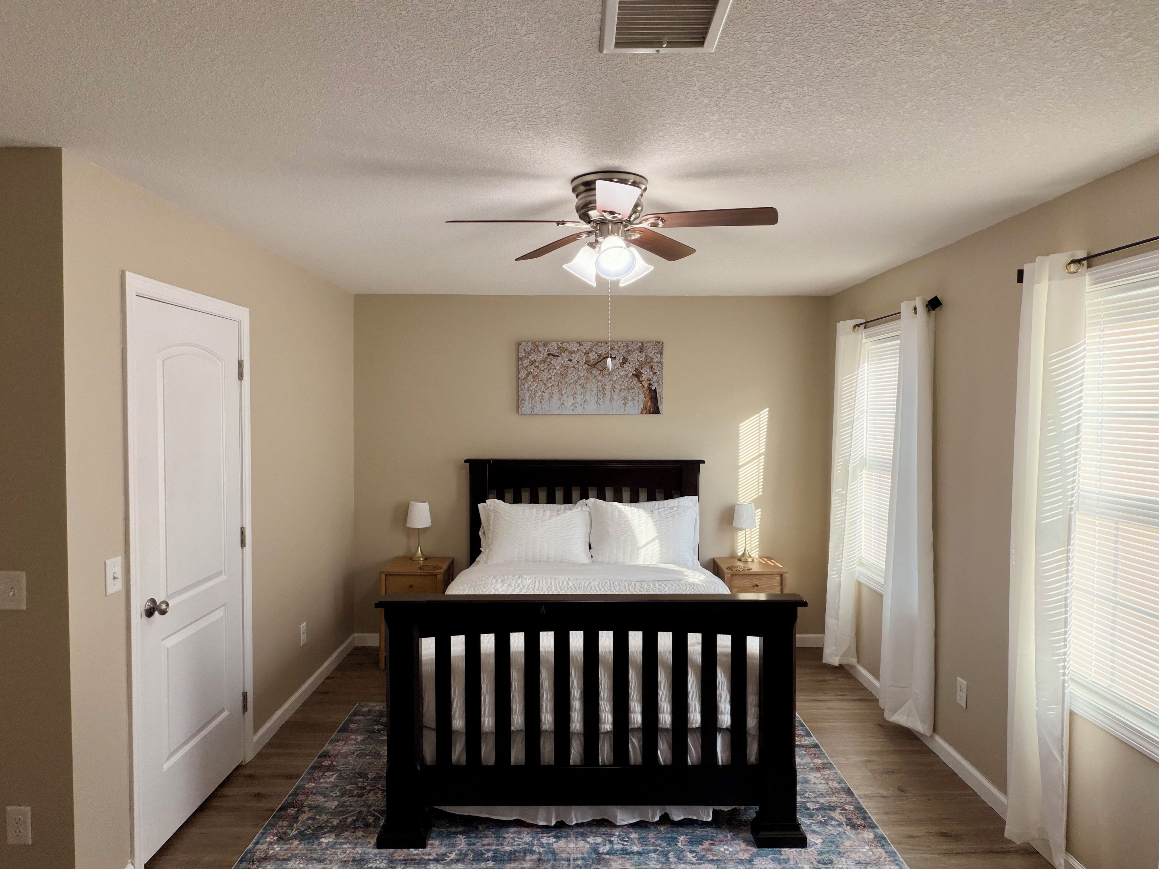 A bedroom with a bed, a ceiling fan, a nightstand with a lamp, a door, and a window with blinds.