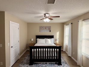 A bedroom with a bed, a ceiling fan, a nightstand with a lamp, a door, and a window with blinds.