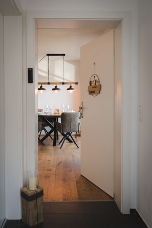 Un couloir mène à une salle à manger où se trouvent une table et des chaises en bois, un luminaire suspendu et un panneau fixé au mur.