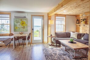 A cozy living room with a wooden ceiling, a sofa, a dining table with chairs, and a view through a glass door. - Redshore River Cottage -Private Waterfront Retreat with Hot Tub (Lot 35)