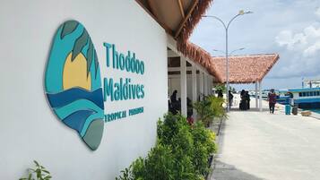 A sign for Thoddoo Maldives with a tropical paradise theme, a covered walkway, and a view of the sea.