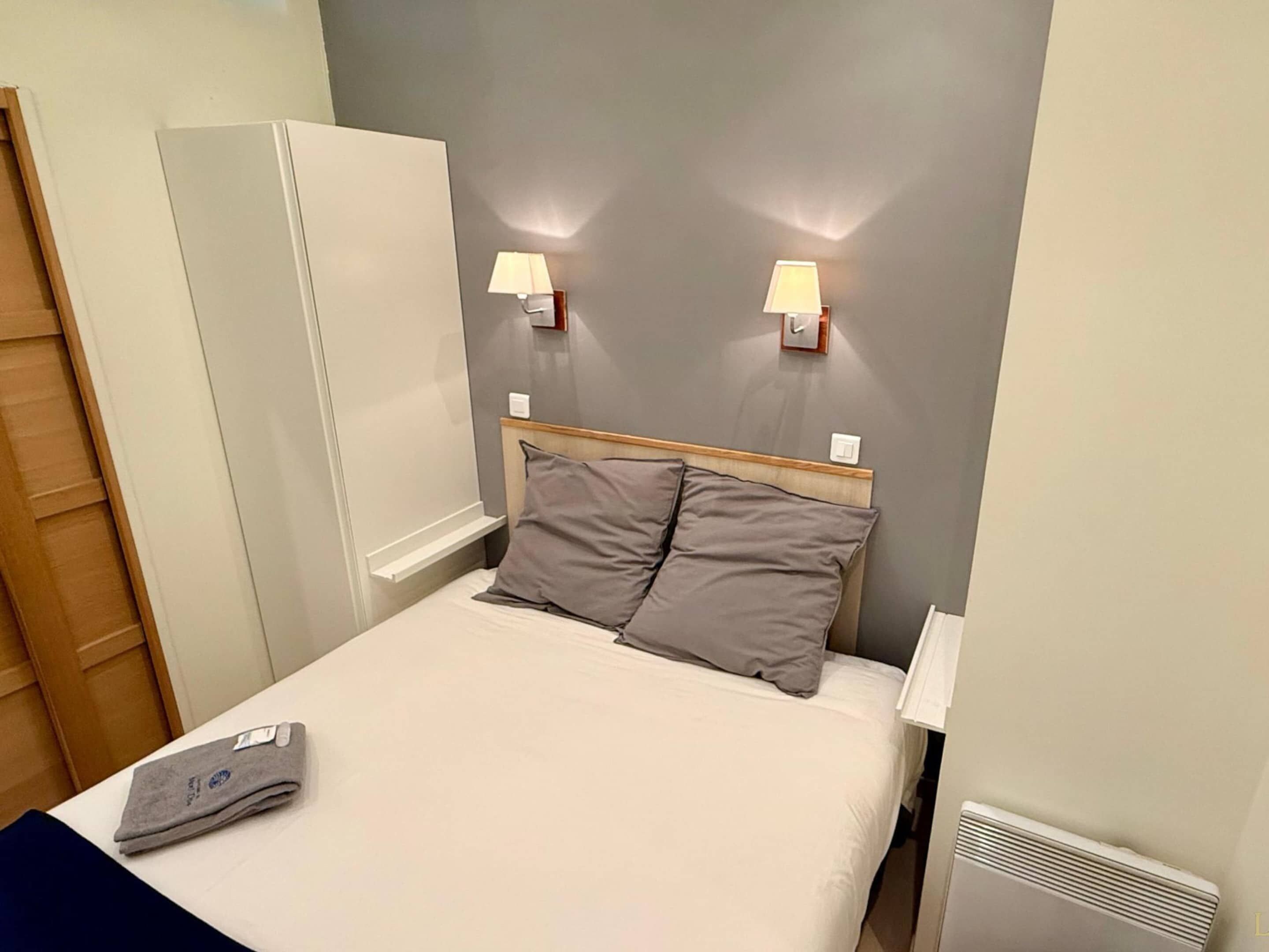 A hotel room with a bed, two bedside lamps, a heater, and a folded towel on the bed.