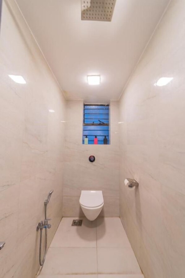 A narrow shower stall with a toilet, a showerhead, and a small window with blue and red elements.