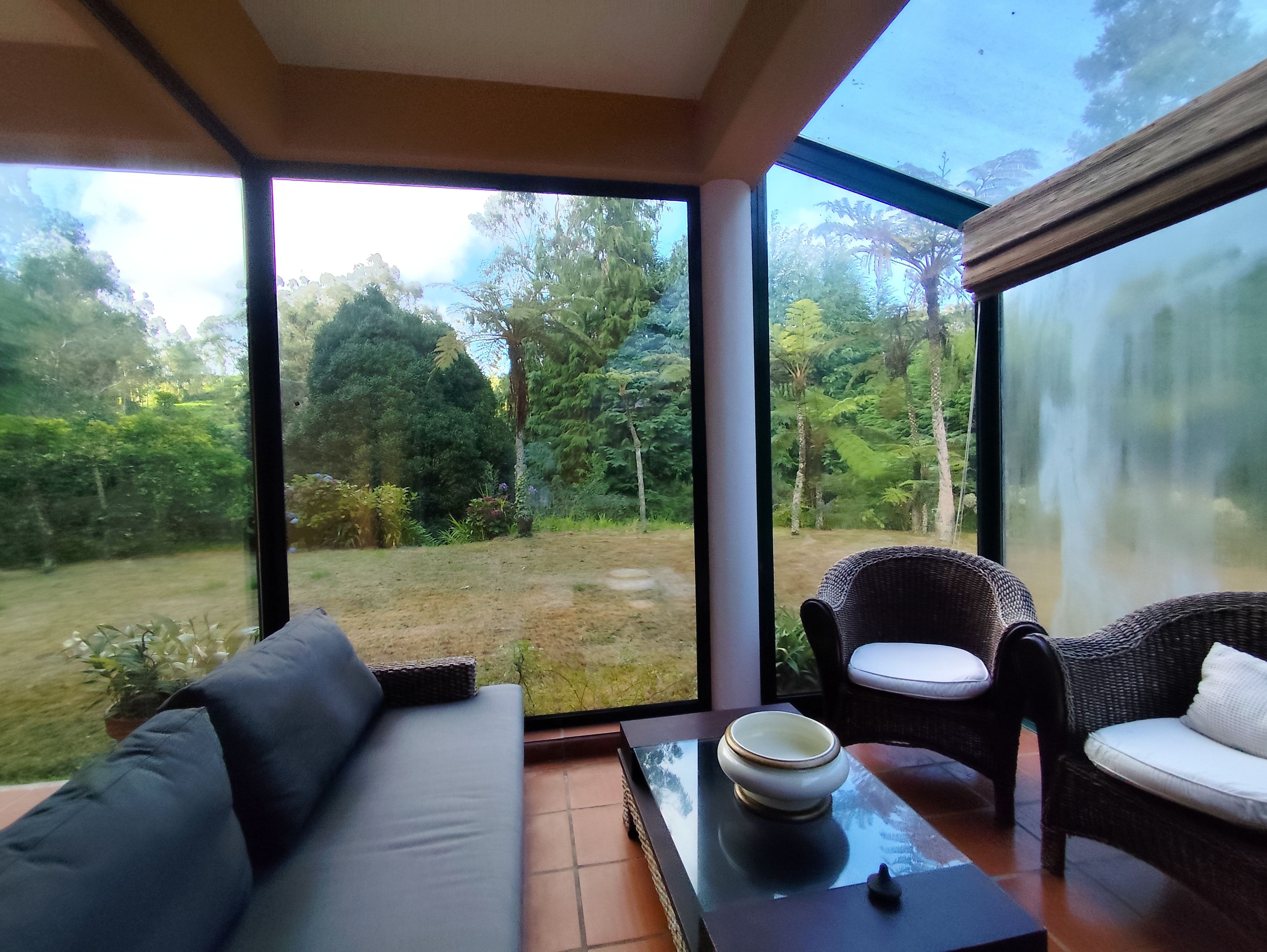 Una habitación con un ventanal grande, un sofá, un sillón de mimbre y una mesa con un cuenco.