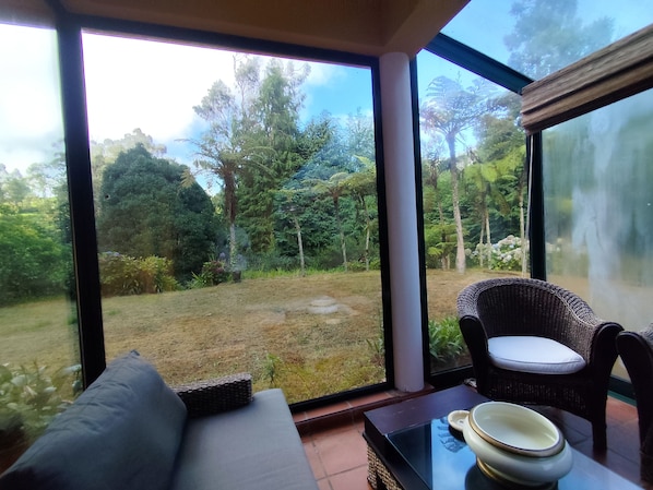 A room with a large window, a wicker chair, a sofa, and a table with a bowl.