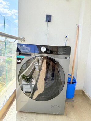 Un lave-linge à chargement frontal doté d’un panneau de commande numérique, d’une prise de courant située au-dessus et d’un seau bleu placé à droite.